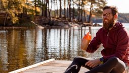 A man is sitting on an exercise mat outdoors by a lake. He is holding a protein shake.